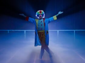 1 clown standing in the middle of the ice skating rink under a spotlight with his arms open wide
