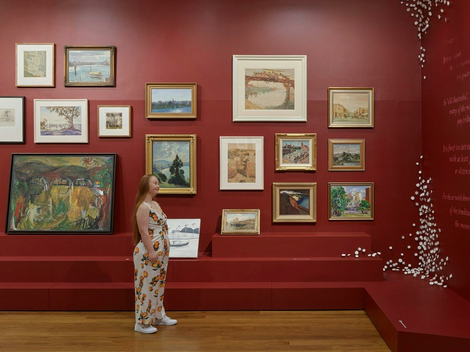 A person in a gallery space with dark red walls looks up at a collection of paintings.