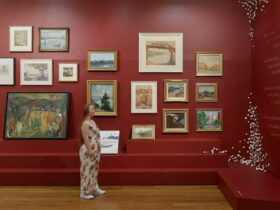 A person in a gallery space with dark red walls looks up at a collection of paintings.