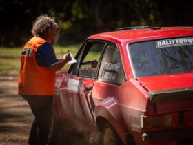 An official in an orange hi-vis vest is talking to two people inside a red rally car
