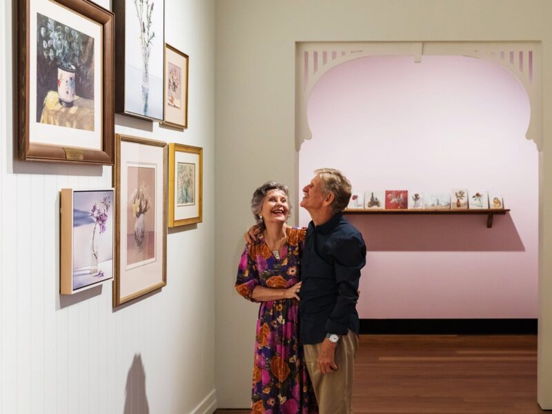 Two people linked arms smiling in a gallery space with paintings hanging on the walls.