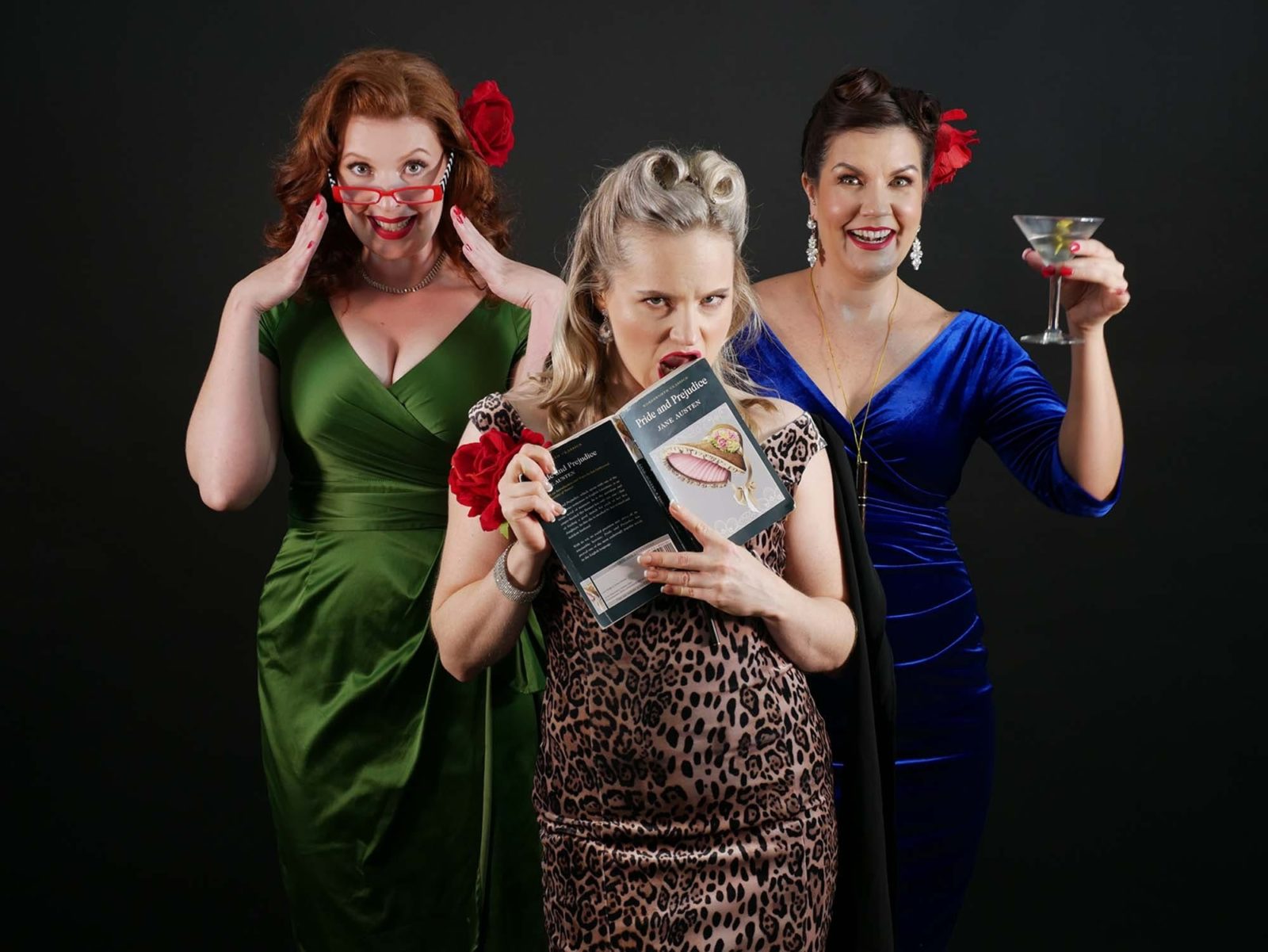 Three people standing against black backdrop, one holds a book, all wearing dresses and red lipstick