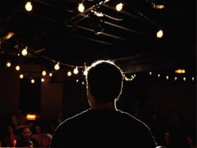 Photo taken from behind of a comedian on stage, their figure silhouetted in front of an audience.