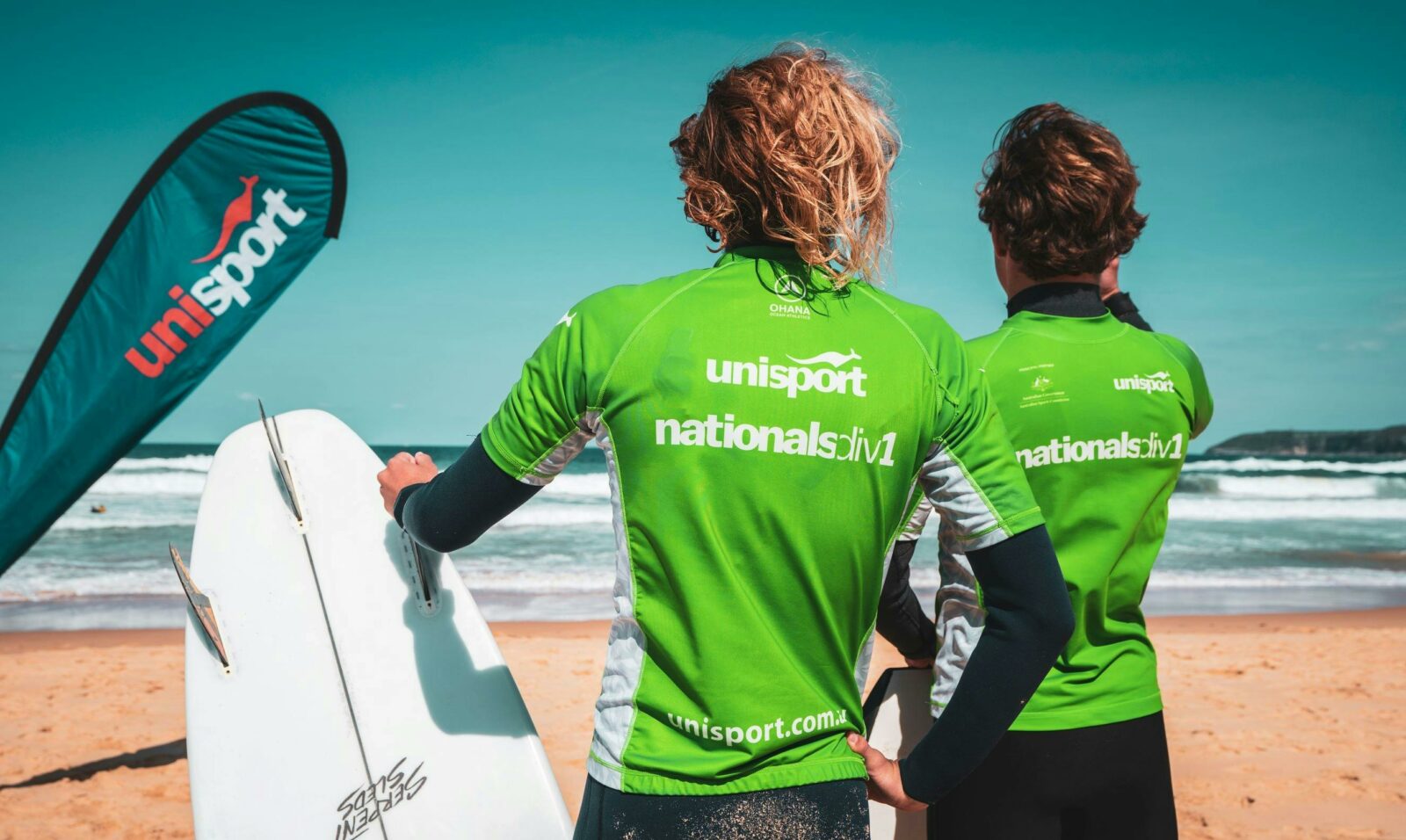 Two male surfers wearing green UniSport rash vests looking at ocean holding surfboard.