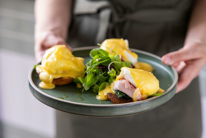 A cropped photo of waiter's hands holding a plate of eggs benedict. Three eggs on muffins with sauce