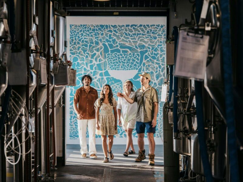 Four people framed by Stainless Steel tanks, in front of a blue and white background