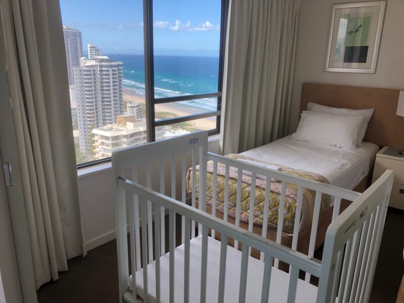 A white cot set up in a hotel room overlooking the beach and Surfers Paradise skyline.
