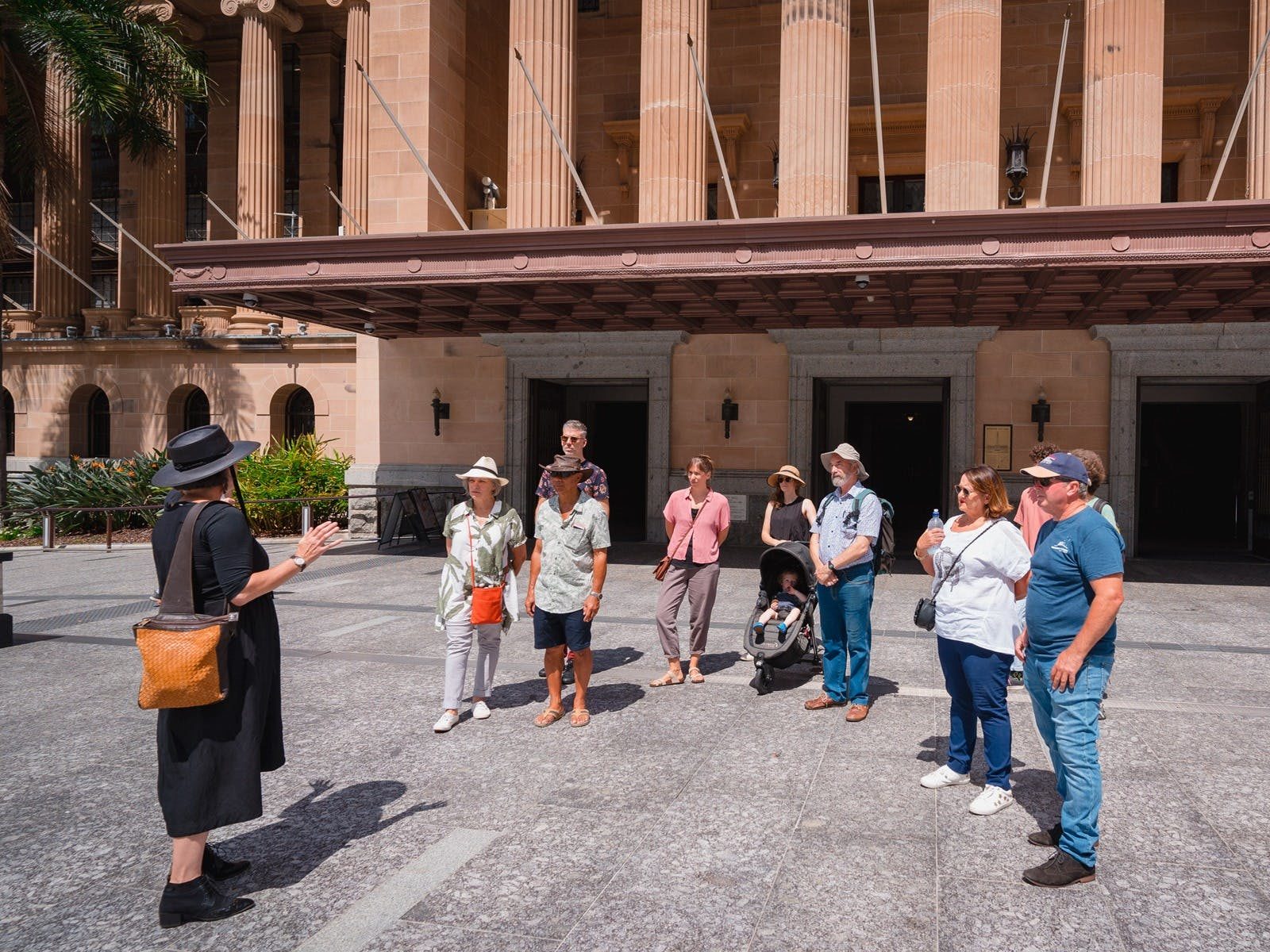 A group of people stand listening to a Museum of Brisbane tour guide in front of City Hall.