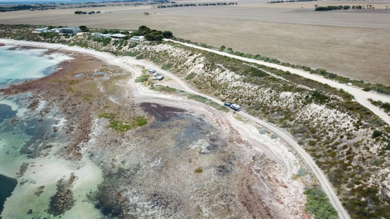 Drone view of campground alongside reef and shallow water. Shacks on clifftop in background.