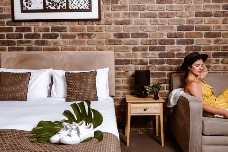 A lady lays on a couch next to a queen-sized bed, which is against a brick wall
