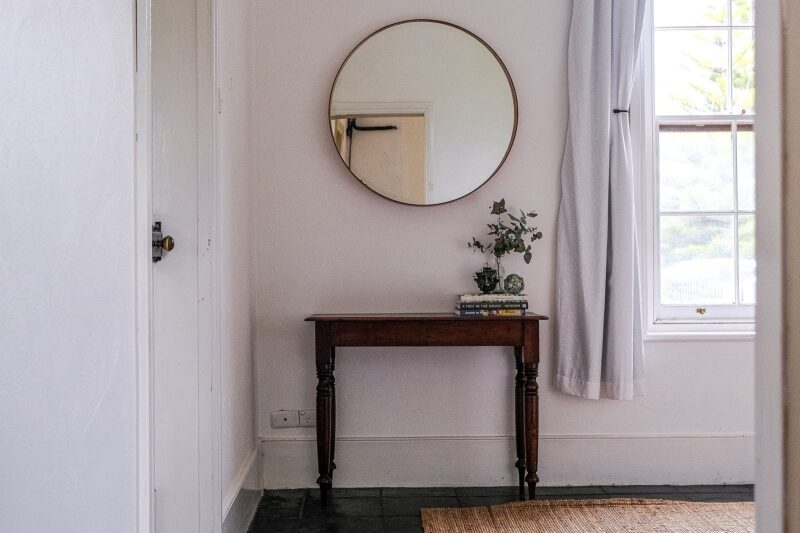 A wooden credenza sits against a wall underneath a round mirror, to the left of a window