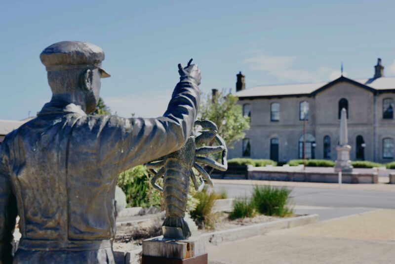 A bronze sculpture of a lobster fishermen against the backdrop of Port MacDonell's Customs House