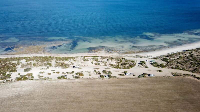 Drone view of sandy tracks forming grid-like campsites. Beach and blue ocean in background.
