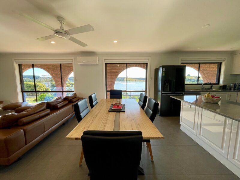 Dining table at centre, lounge to left and kitchen to the right with 3 large windows with lake views