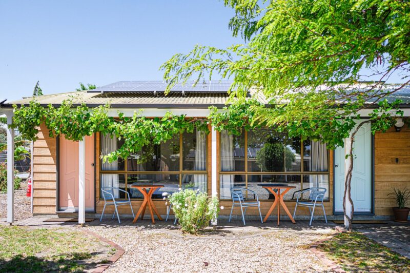 Exterior of rooms, pastel pink and blue doors, outdoor table and blue chairs, grape vine trellis.