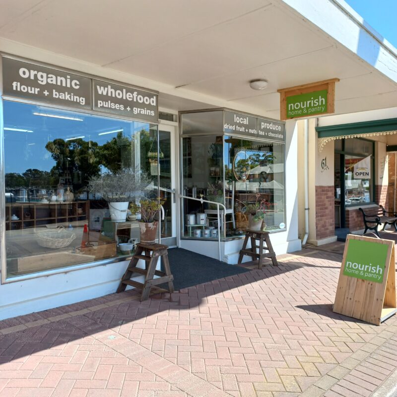 Image shows the mostly glass shop front of Nourish Home & Pantry with planters and bright green logo