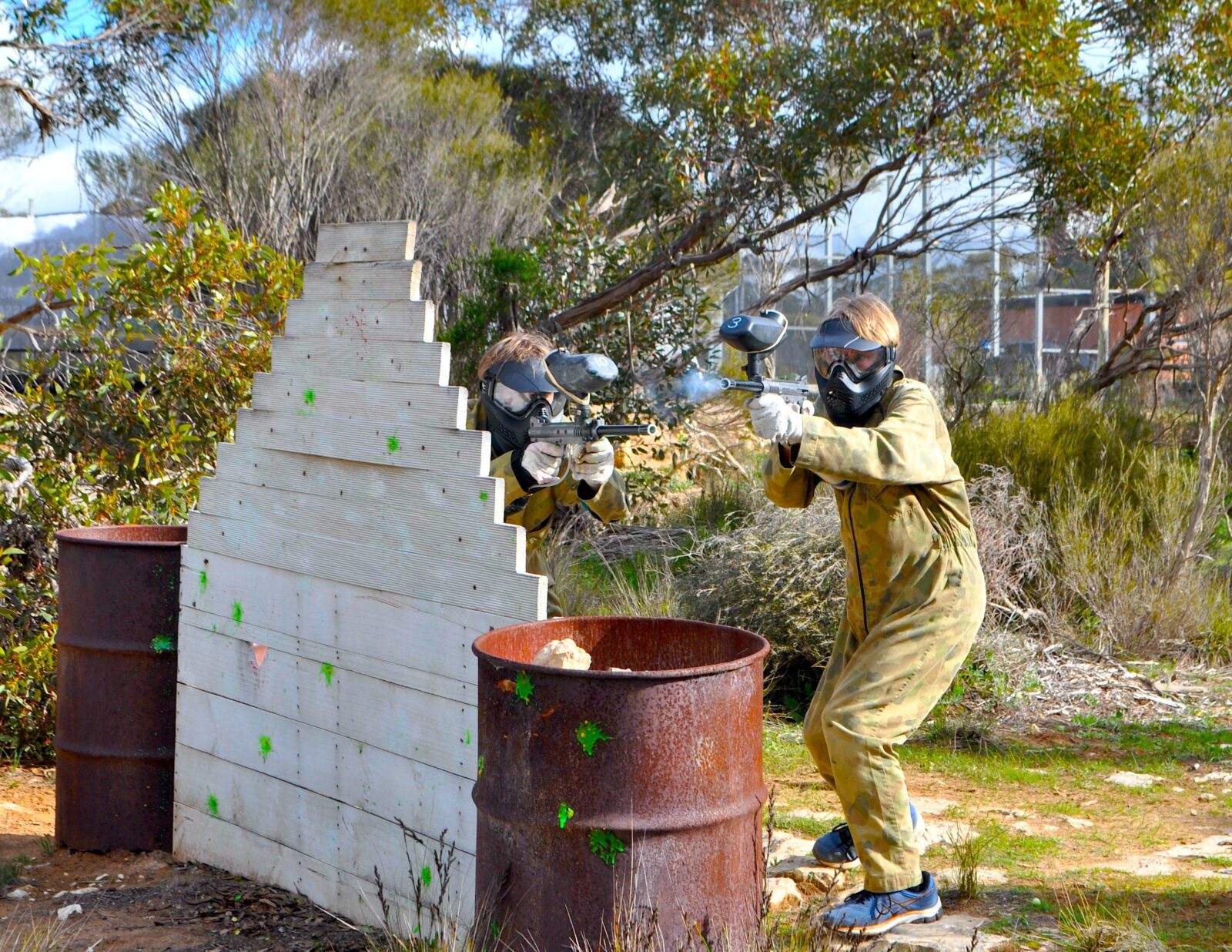 Action shot of two paintball players hiding behind a barricade in a rural bush setting.