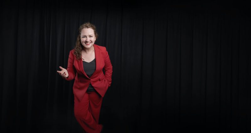KC wears a striking red pants suit and black top. She is smiling in front of a black stage curtain.