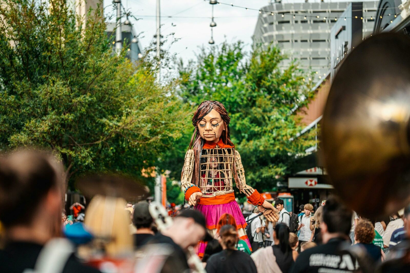 A 3.5m tall puppet of a 10 year old girl is surrounded by a crowd. She has her eyes closed.