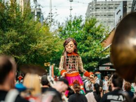 A 3.5m tall puppet of a 10 year old girl is surrounded by a crowd. She has her eyes closed.