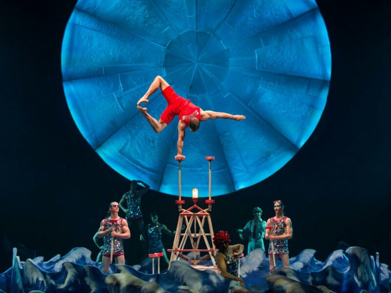 A performer wearing bright red, balances on one hand in front of a bright blue circle backdrop.
