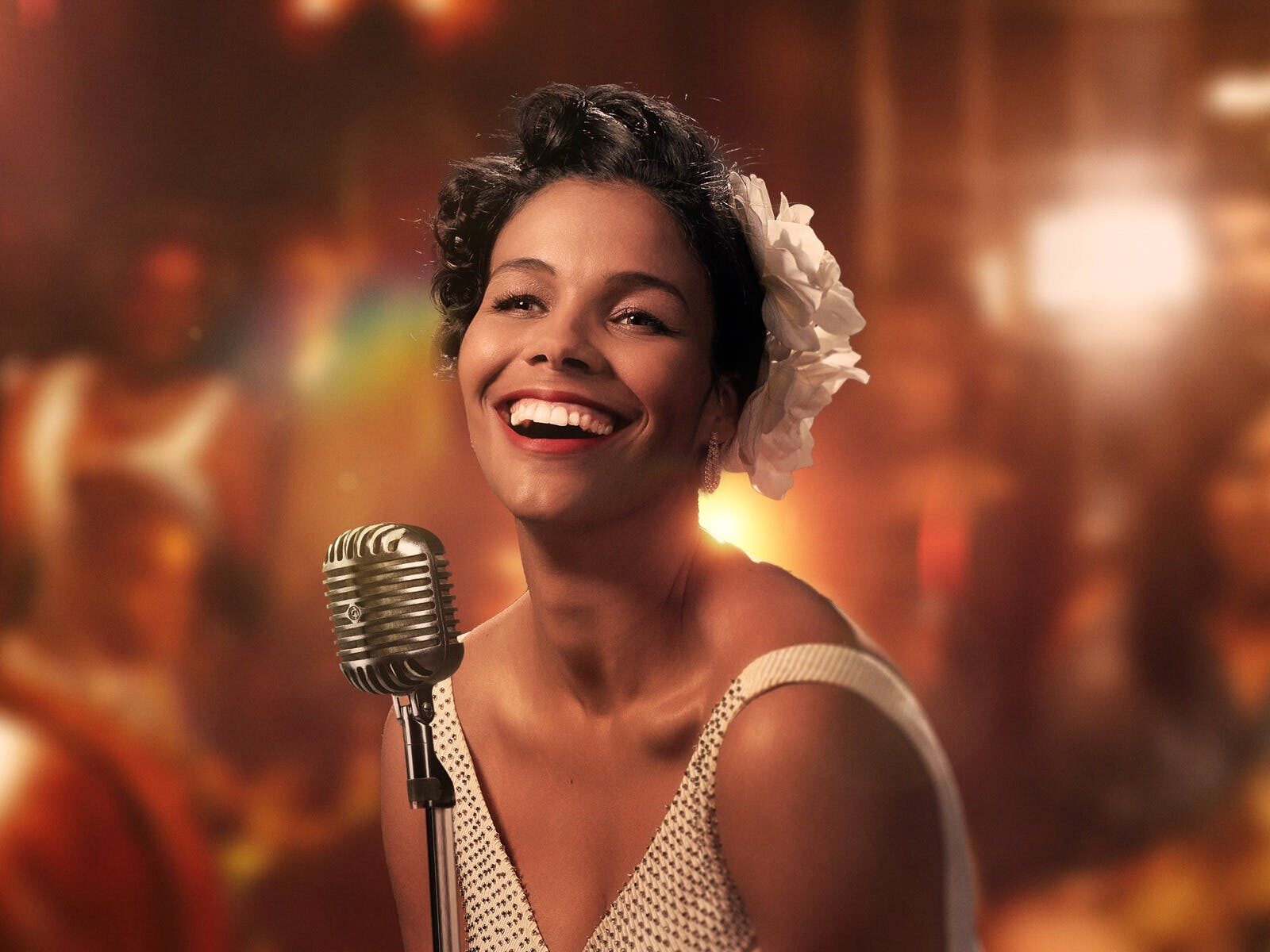 Zahra Newman wears a gardenia in her hair and smiled at the camera, stands in front of a vintage mic
