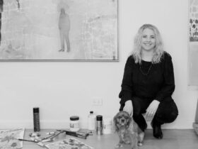 Artist Belinda Wilson crouches with dog on studio wall, paints on floor, paintings on either side