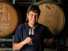 Male winemaker in navy polo shirt smiling and holding a glass of red wine in front of wine barrels