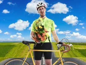 A man in front of a scenic field and sky holding flowers, a bicycle, wearing helmet