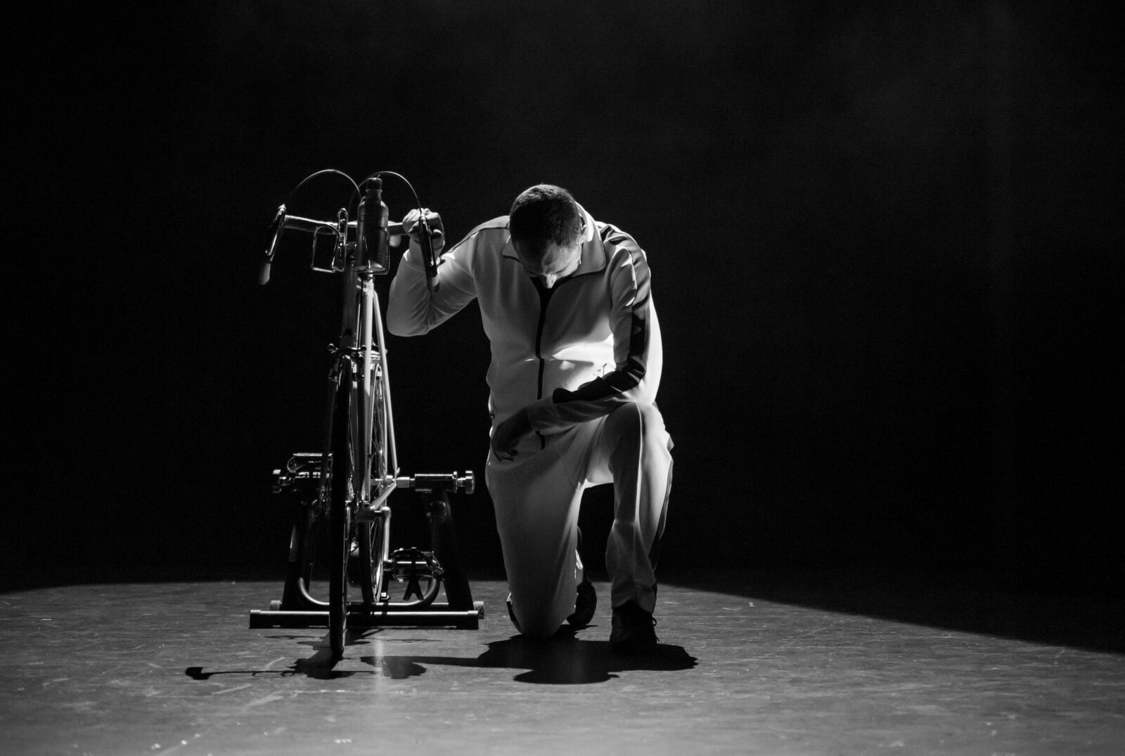 A man in a white tracksuit kneels beside a bicycle, the photo is black and white.