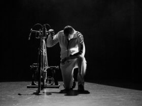 A man in a white tracksuit kneels beside a bicycle, the photo is black and white.