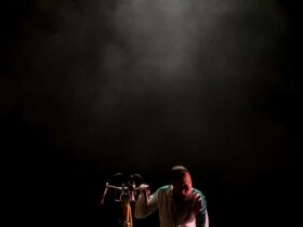 A man in a white tracksuit kneeling next to a bicycle on a black stage under a spotlight