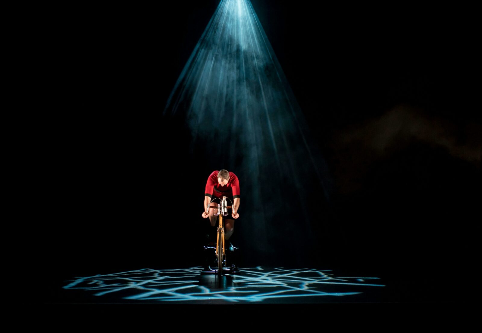 A man lit by a blue textured spotlight is riding a bicycle on stage wearing a red riding suit.