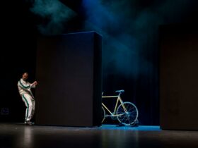 A man in white pulls on a large black cube to reveal a bicycle illuminated by a blue spotlight