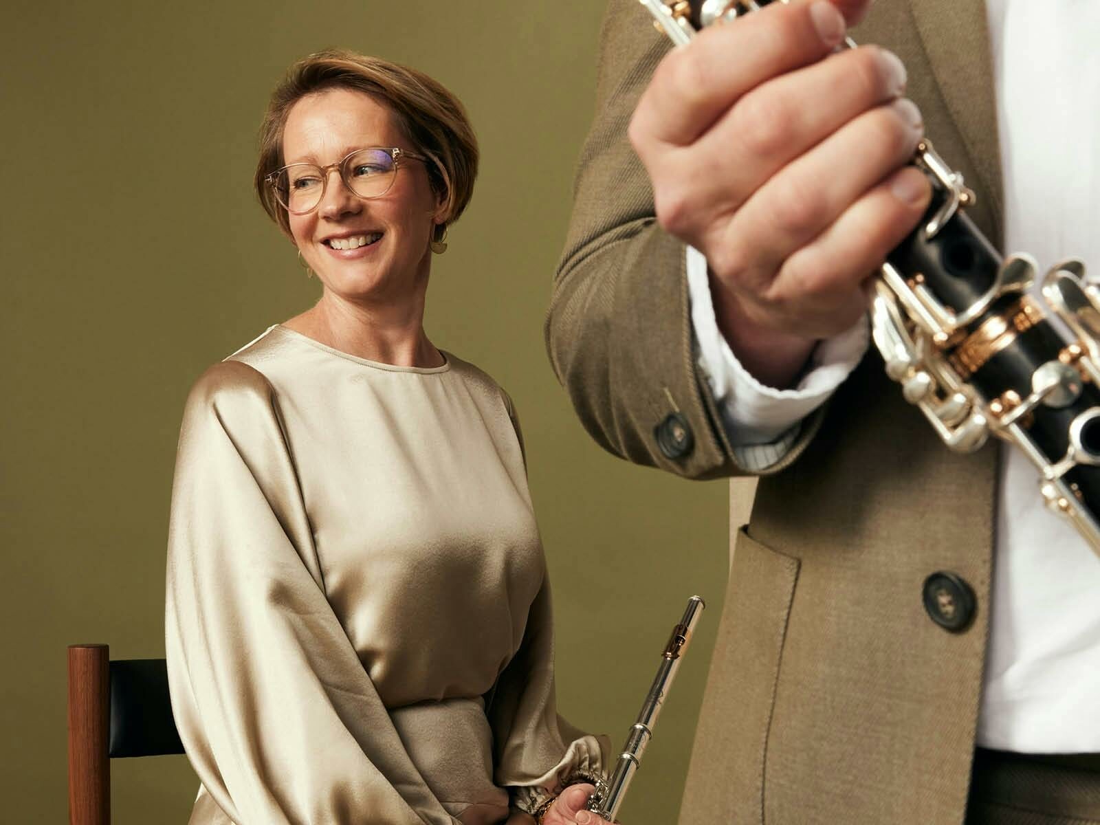 A woman in a Champane colour dress, sits, holding a flute with part of a clarinet and hand visable