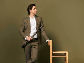 A man in a khaki suit and white shirt stands holding a clarinet looking off to the side.