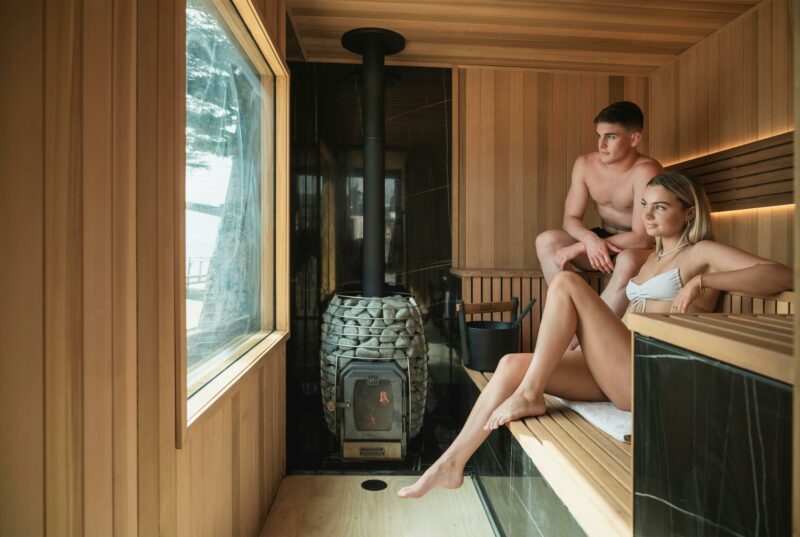 A male and female sit comfortably in the timber clad sauna looking out the viewing window to the sea