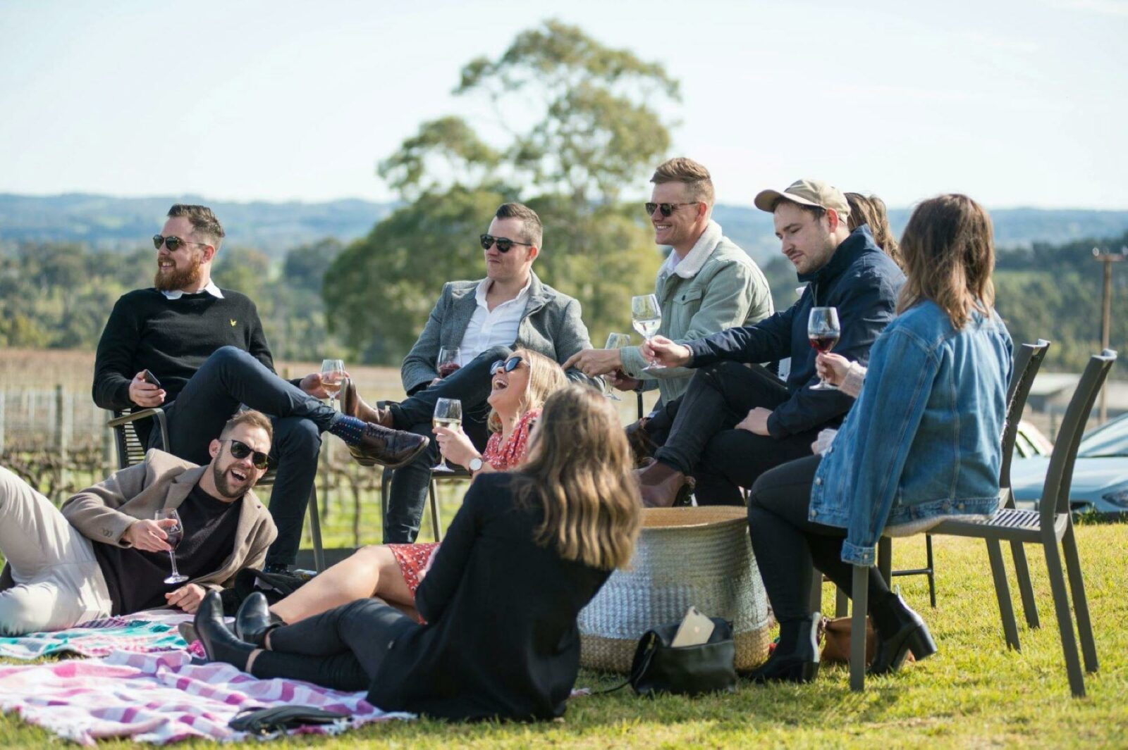 A group of friends sitting on chairs and a picnic rug , enjoying a glass of wine and the views