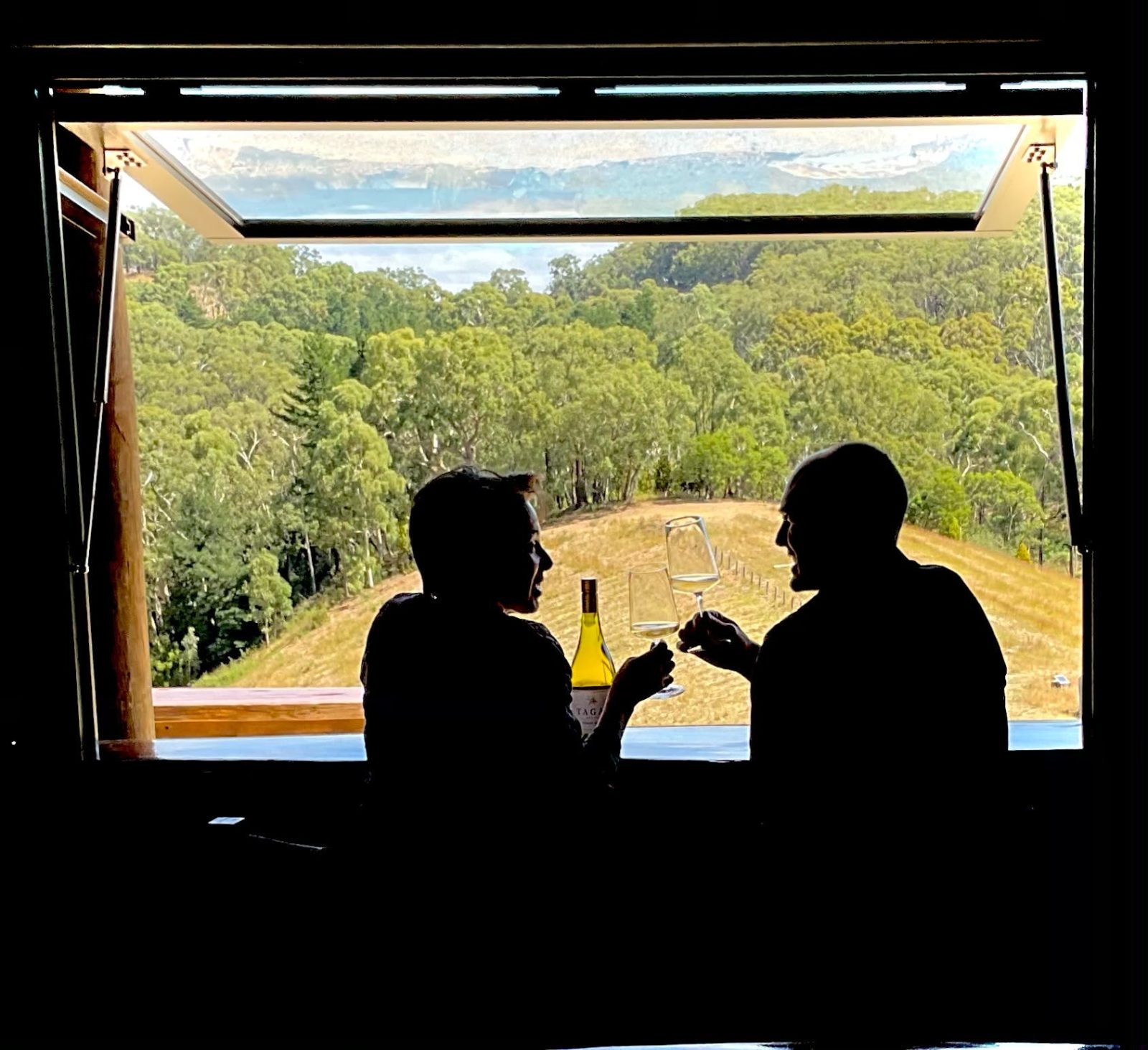 a couple sit in front of a view of Australian bushland drinking a glass of wine