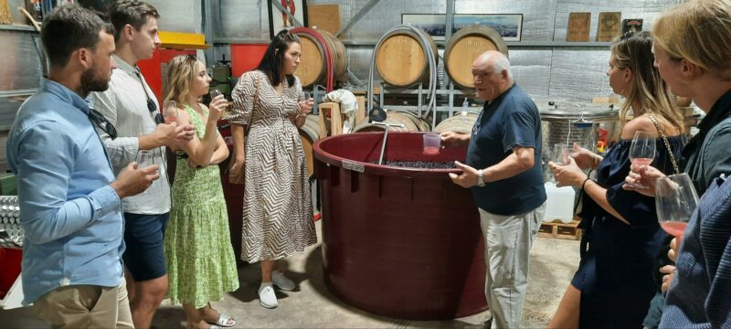 A group of 6 guests and the winemaker in a shed standing around waiting to have glassed filled.