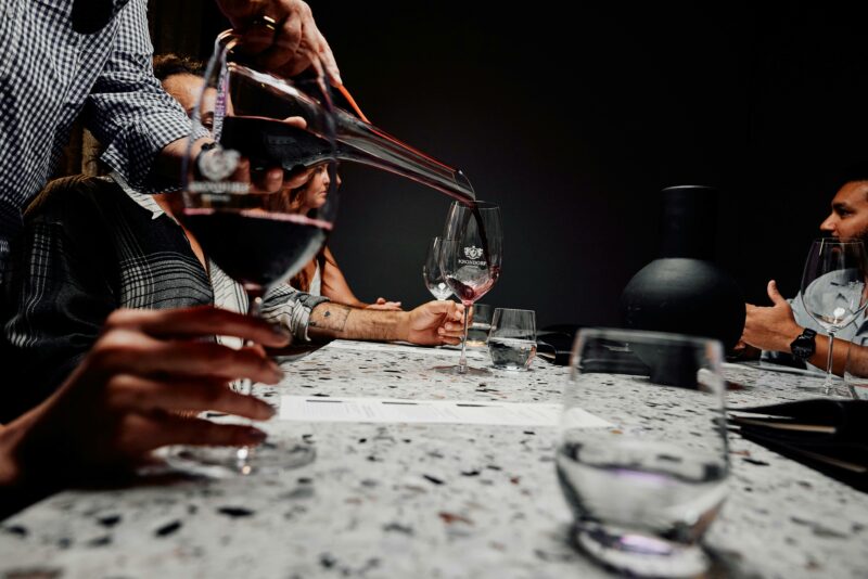 A view of a table showing red wine in stemmed glasses and wine being poured from a decanter