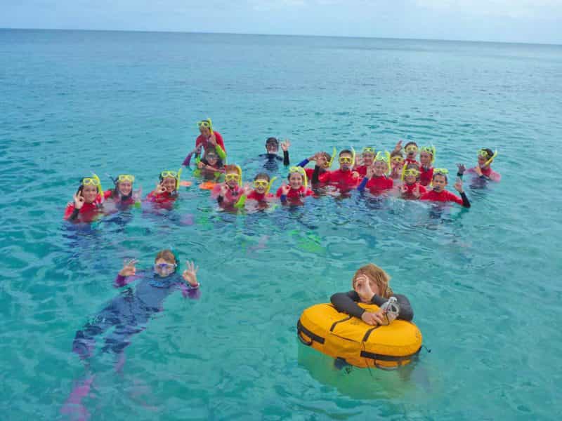 A large group of kids and instructors smile and wave in snorkels