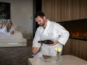 A man pouring a glass of champagne by the fireplace, a woman laying on the bed is smiling