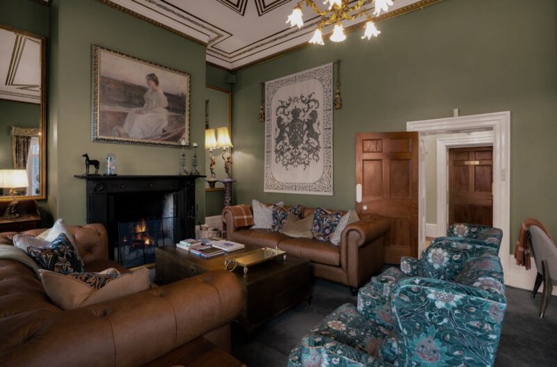 Green painted room filled with plush furniture in front of a roaring open fire place