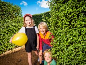 Three children in a lush green hedge maze at Amaze Richmond. Girl is holding a yellow balloon.