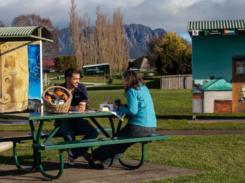 Man and Woman sit at a picnic table in Mural Park, with a picnic lunch. with Mt Roland