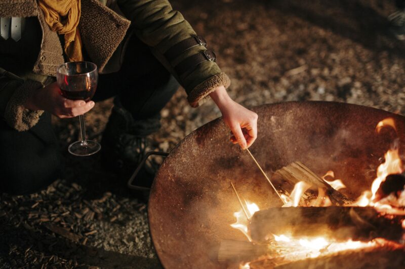 A marshmallow being toasted over a campfire while the individual holding a glass of red wine