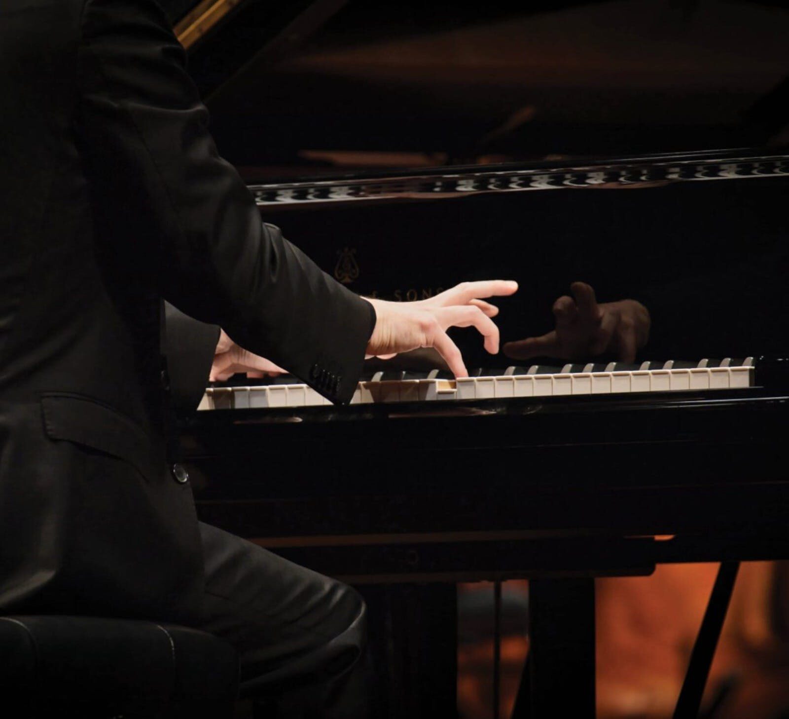A musician in a black suit, whose back is facing the camera, sitting at a Steinway grand piano.