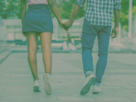 two people holding hands walking down an empty road, one wears a skirt, the other jeans