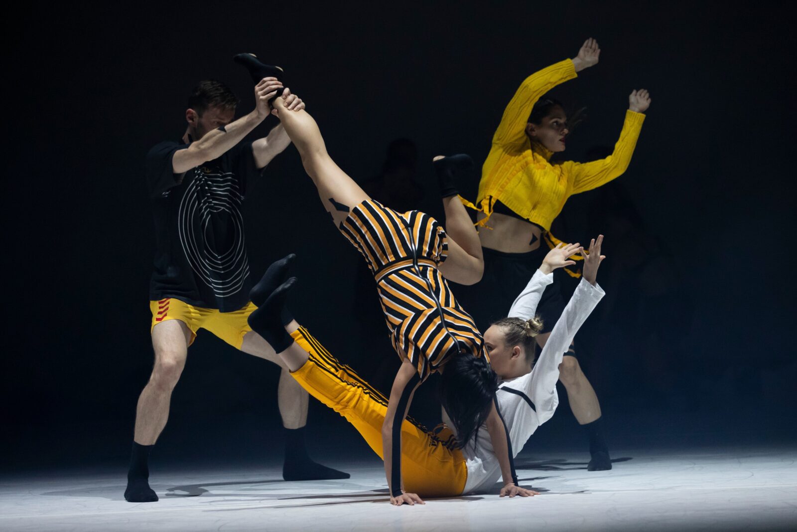 4 dancers pose in a group. One sits with hands and legs in the air. One does a handstand. Two stand.